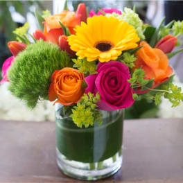 Bright bouquet of yellow gerbera, orange and pink roses, and green blooms in a clear glass vase