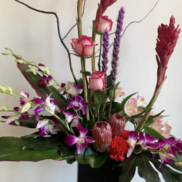 Tall floral arrangement with pink roses, purple orchids, and red tropical blooms in a black vase