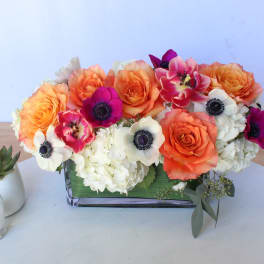 Orange and white floral arrangement in a rectangular glass vase beside a small succulent and candle