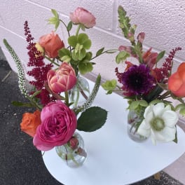 Three small mixed flower arrangements in clear glass vases on a white table.
