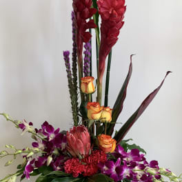 Tall tropical arrangement with red blooms, orange roses, and purple orchids in a wooden container