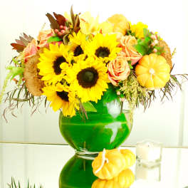 Sunflower and peach rose arrangement in a green glass vase with small pumpkins