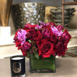 Red and pink rose bouquet in a square glass vase with a black candle beside it