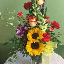Tall mixed bouquet with sunflowers, roses, orchids, and carnations in a glass vase