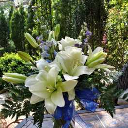 White lily and rose arrangement with blue ribbon and purple accents in a clear vase on an outdoor table