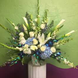 Tall floral arrangement with white lilies, roses, and blue hydrangeas in a white pedestal vase