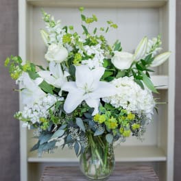 Tall arrangement of white lilies, roses, hydrangeas, and yellow-green blooms in a clear glass vase