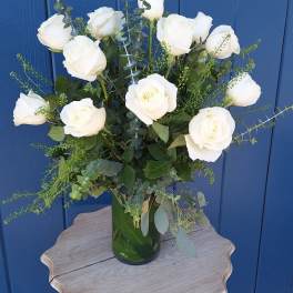 Tall arrangement of white roses with filler in a clear glass vase on a small wooden table