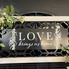 Framed sign with white floral accents and the words "LOVE brings us home"