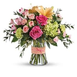 Mixed bouquet of pink roses, lilies, and green hydrangeas in a clear vase with a peach ribbon.