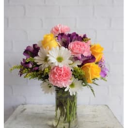 Yellow roses, pink carnations, white daisies, and purple blooms in a clear glass vase