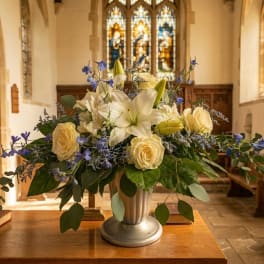 Arrangement of white lilies, cream roses, and blue flowers in a silver urn on a wooden table in a church.
