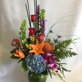 Tall mixed flower arrangement in a green glass vase with lilies, roses, orchids, and hydrangea