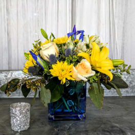 Compact arrangement of yellow lilies, cream roses, and blue accents in a blue glass cube vase.