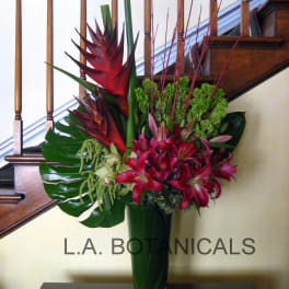 Tropical flower arrangement in a tall glass vase with red lilies and green foliage