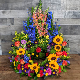 Tall circular wreath with bright sunflowers, roses, carnations, and blue delphinium.