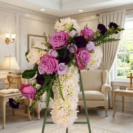 Tall cross-shaped spray of white mums with pink and purple roses on a metal easel indoors.