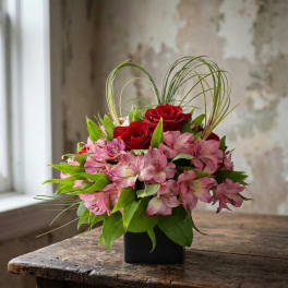Compact arrangement of red roses and pink alstroemeria in a black cube vase with looped grass forming a heart shape.
