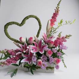 Pink and lavender floral arrangement with a moss heart frame in a white container