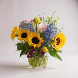 Mixed bouquet with sunflowers, blue flowers, and pink blooms in a glass vase