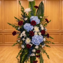 Tall standing spray with blue hydrangeas, white mums, red carnations, and large green leaves on an easel.