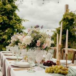 Pastel rose centerpiece on an outdoor banquet table with crystal goblets, taper candles, and grapes on a green runner.