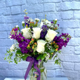 White roses and purple flowers arranged in a glass vase with a purple ribbon.
