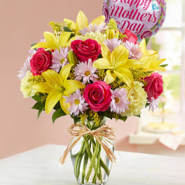 Mixed bouquet of yellow lilies, pink roses, and pastel daisies in a glass vase with a Mother’s Day balloon.