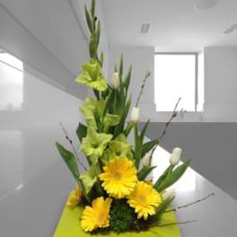 Tall floral arrangement with yellow gerbera daisies and green lilies on a square base