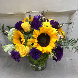 Bouquet of sunflowers, yellow roses, and purple blooms in a glass vase