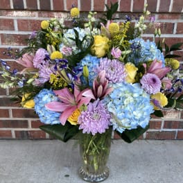 Mixed bouquet of blue hydrangeas, pink lilies, and yellow roses in a glass vase