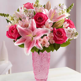 Arrangement of pink lilies and roses with white accent flowers in a tall pink glass vase