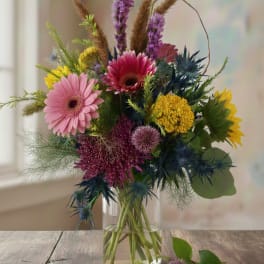 Tall mixed bouquet with pink gerbera daisies, yellow blooms, and purple accents in a clear glass vase