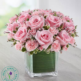 Compact arrangement of pink roses and carnations in a clear square vase lined with green leaves