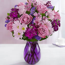 Mixed bouquet of pink roses, daisies, and purple blooms arranged in a purple glass vase