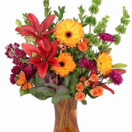 Tall arrangement of orange gerberas, red lilies, and mixed blooms in an amber glass vase.