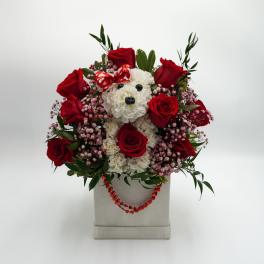 Red roses and white flowers arranged around a teddy bear in a box