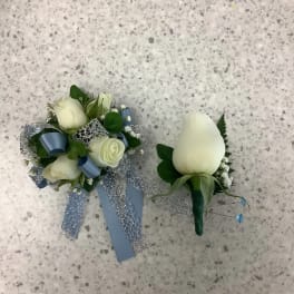 Two white rose boutonnieres with blue ribbon accents on a speckled floor