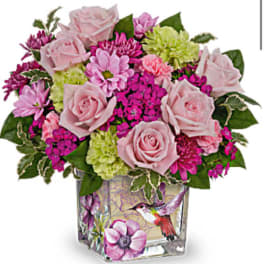 Compact arrangement of pink roses and bright blooms in a square glass vase with a hummingbird design
