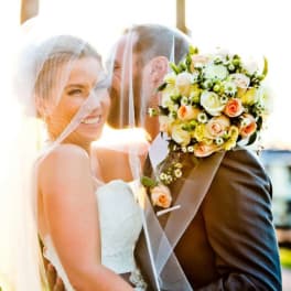Bride and groom under a veil, holding a bouquet of pale roses