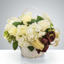 White roses and hydrangeas in a white wicker basket with a gold and burgundy ribbon bow
