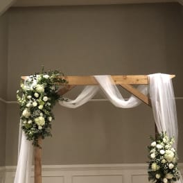 White floral arrangements on a wooden wedding arch with draped white fabric