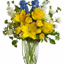 Yellow roses and lilies with blue and white flowers in a clear vase