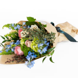 Wrapped bouquet of pink and cream roses with blue accent flowers in kraft paper tied with a dark ribbon