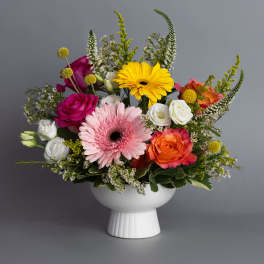 Colorful mix of gerbera daisies, roses, and lisianthus in a white pedestal bowl