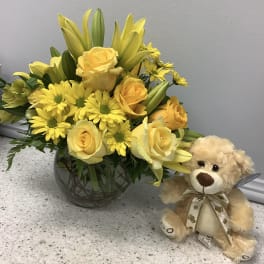Yellow roses and daisies in a glass vase beside a teddy bear