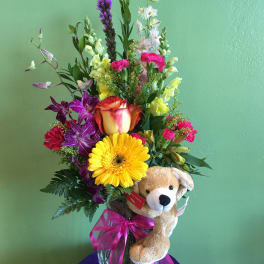 Mixed bouquet in a glass vase with a plush teddy bear and pink ribbon