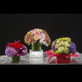 Three colorful floral arrangements in glass and gift boxes on a marble surface