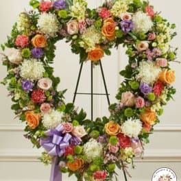 Heart-shaped standing wreath of mixed roses, carnations, and mums in soft colors with a lavender bow
