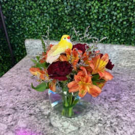 Bouquet of orange and burgundy flowers in a clear glass vase with a yellow bird figurine